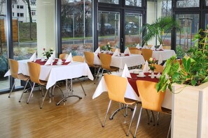 Un restaurant avec des tables dressées pour le service, de grandes fenêtres et des plantes d’intérieur.
