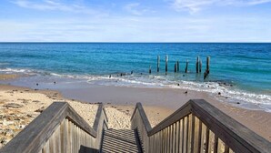 Un escalier en bois mène à une plage où l’eau est d’un bleu cristallin, et des poteaux en bois sont enfoncés dans l’eau.