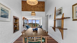 Un coin repas avec une table en bois, des chaises et un luminaire suspendu. Des étagères avec divers objets sont fixées aux murs, et on peut admirer la vue à travers les portes vitrées.