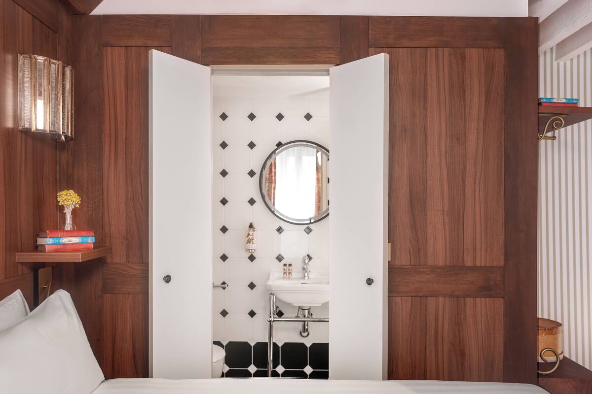 A bedroom with a bed, a bedside table with books, a mirror, and a small bathroom with a sink and toilet.