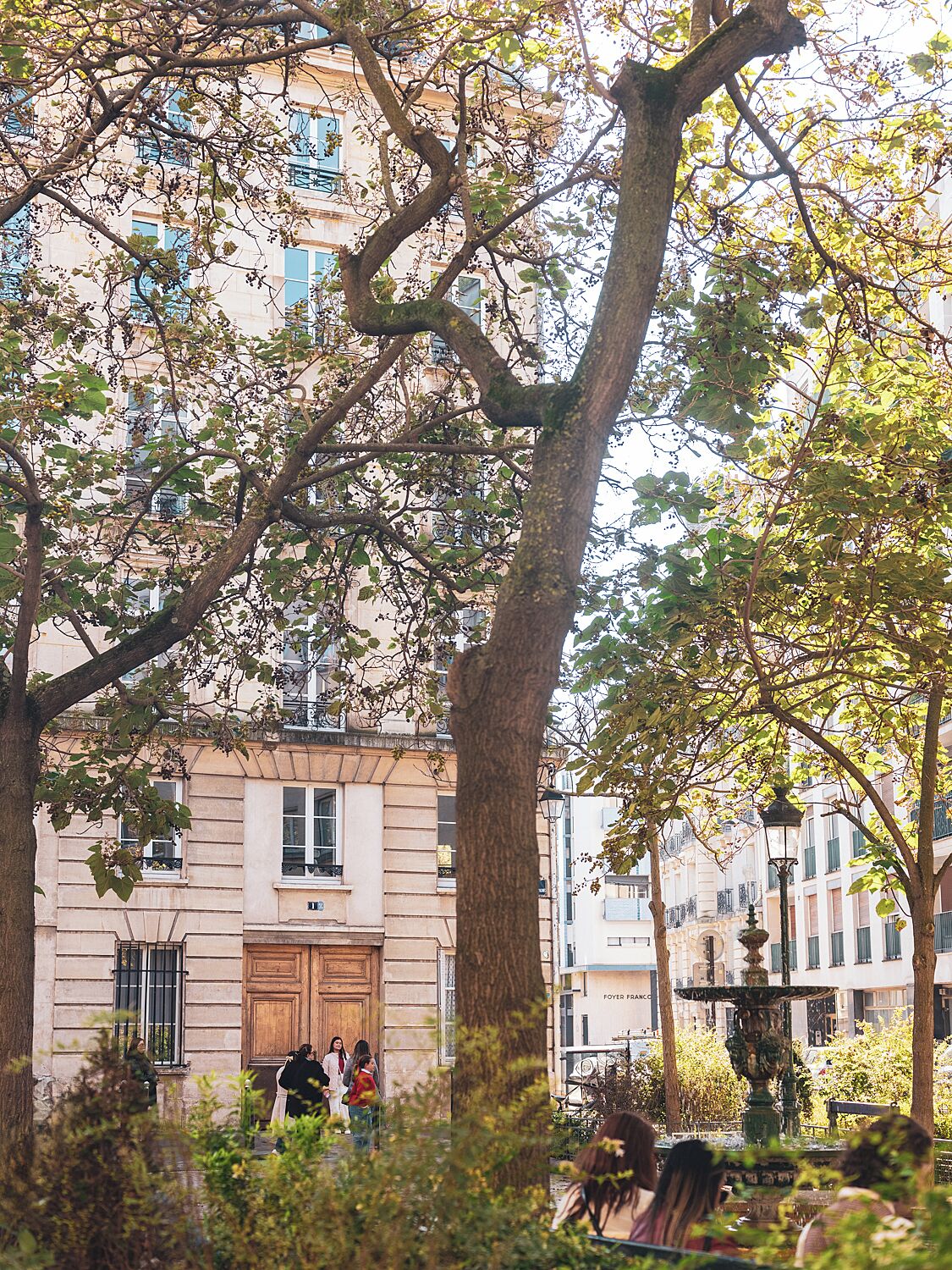 Un edificio con un árbol grande en primer plano, una fuente y gente caminando.