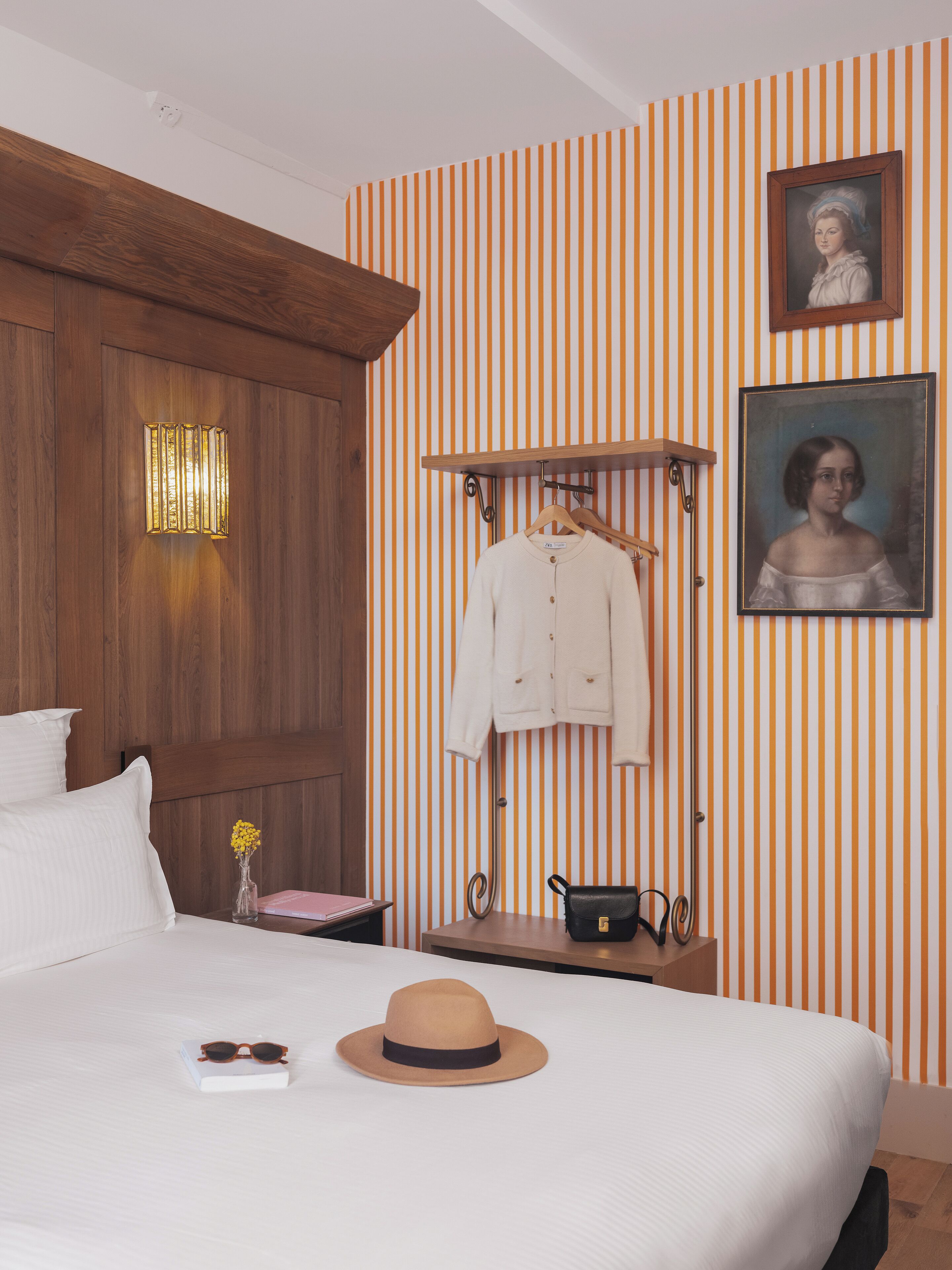 Une chambre d’hôtel avec une tête de lit en bois, un lit avec une literie blanche, un chapeau, un livre, un vase avec des fleurs, un porte-manteau avec un manteau blanc, un sac à main noir et un portrait encadré accroché au mur.