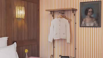 Une chambre d’hôtel avec une tête de lit en bois, un lit avec une literie blanche, un chapeau, un livre, un vase avec des fleurs, un porte-manteau avec un manteau blanc, un sac à main noir et un portrait encadré accroché au mur.