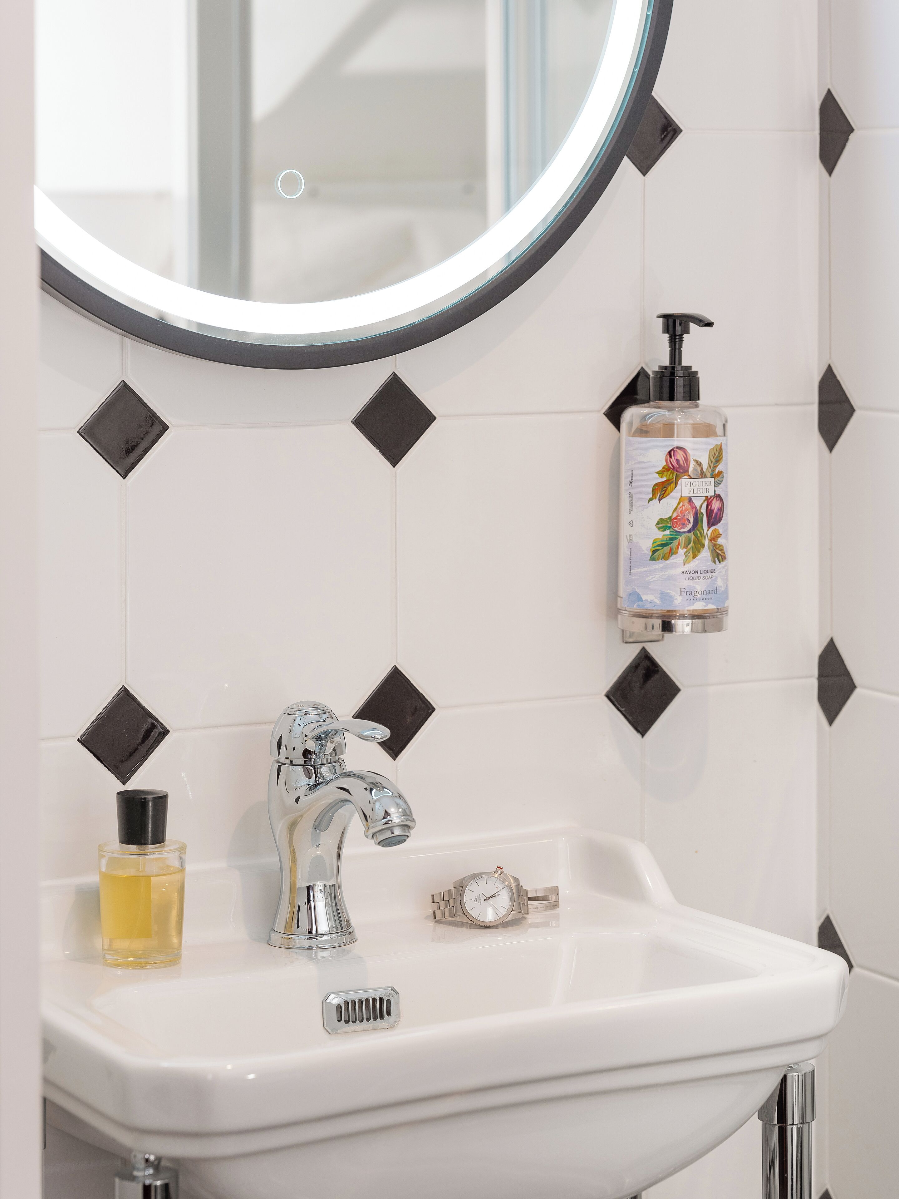 Un baño con un lavamanos blanco, un grifo cromado, un espejo redondo y un dispensador de jabón con diseño floral.