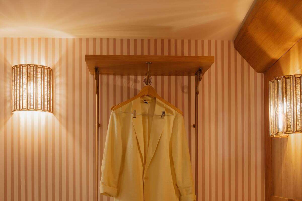 A hotel room with striped wallpaper, a wooden headboard, a hanging coat rack, a bedside table with shoes and a lamp, and a chair.