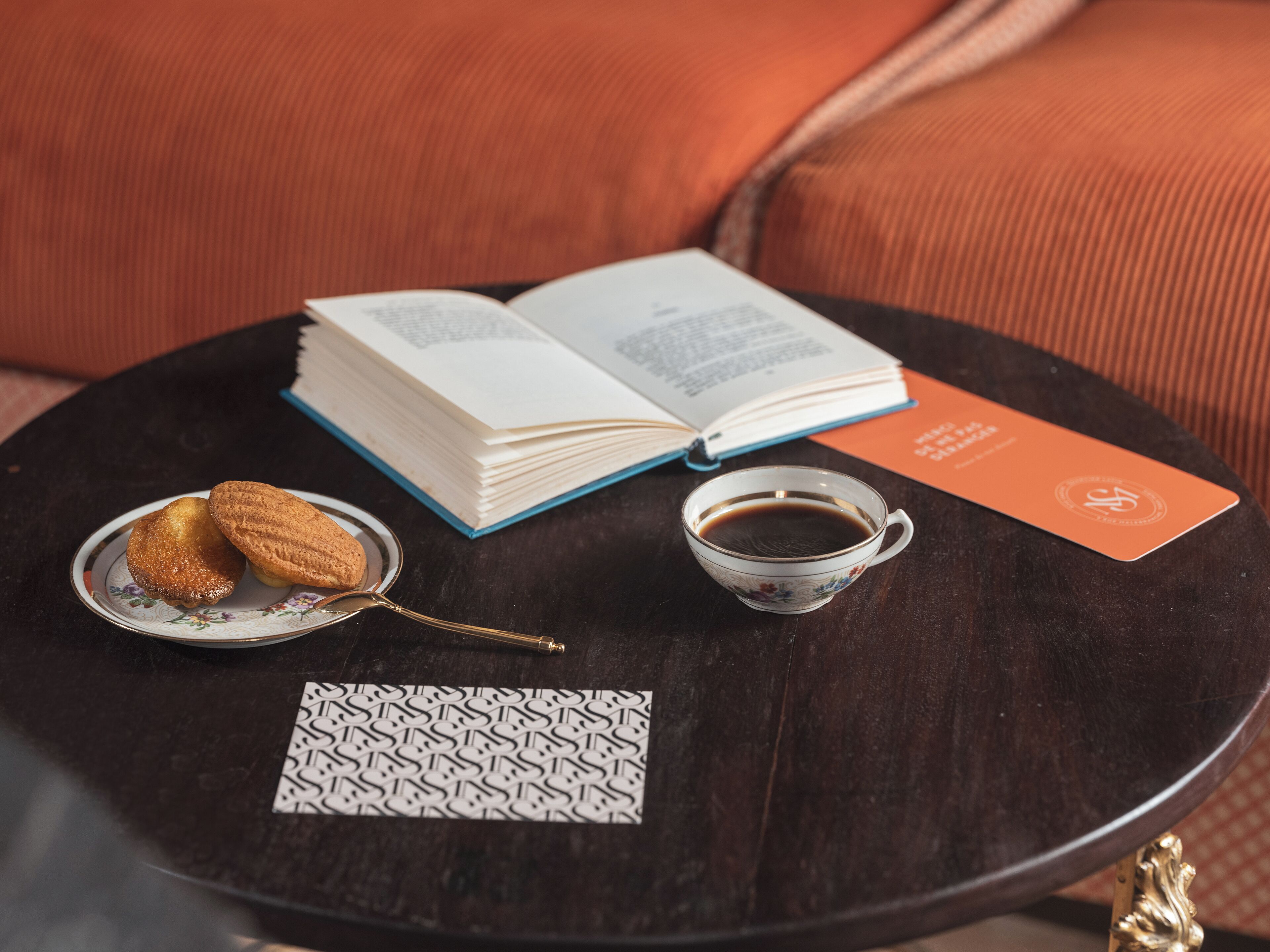 Une table ronde en bois sur laquelle se trouvent un livre, une tasse de café, deux biscuits et une carte de visite.