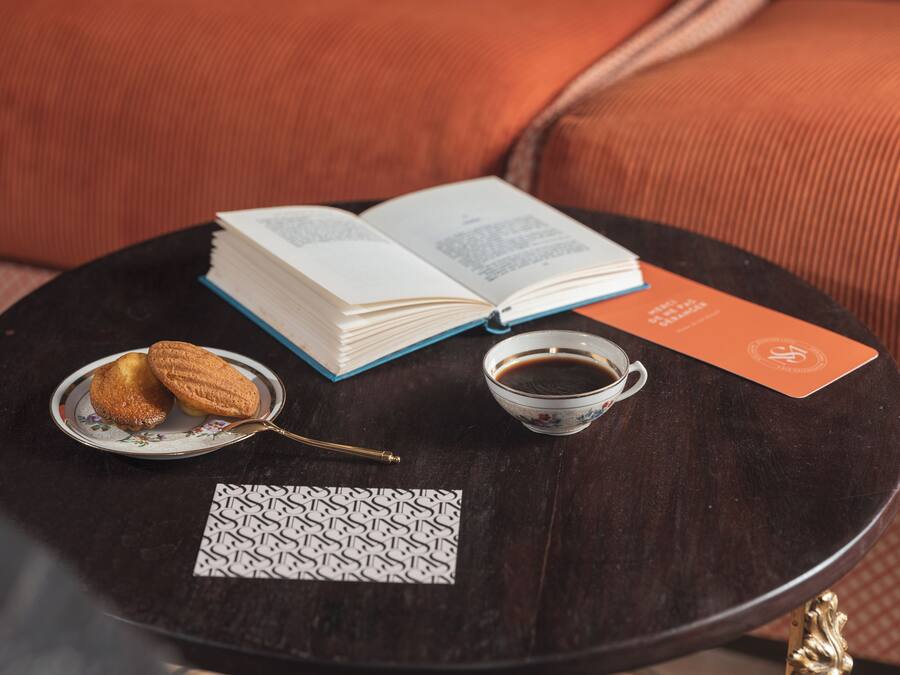 Une table ronde en bois sur laquelle se trouvent un livre, une tasse de café, deux biscuits et une carte de visite.