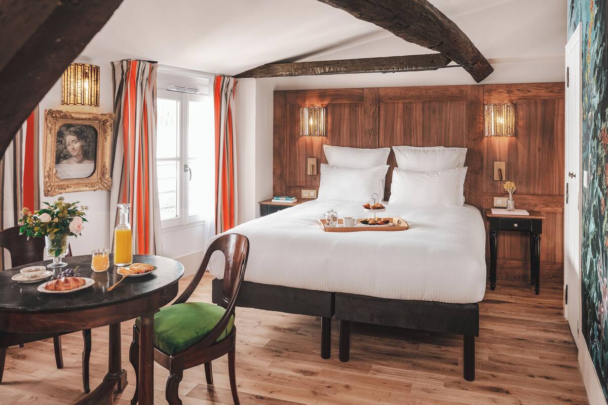 A hotel room with a large bed, wooden ceiling beams, a dining table with breakfast, and a view of the city.