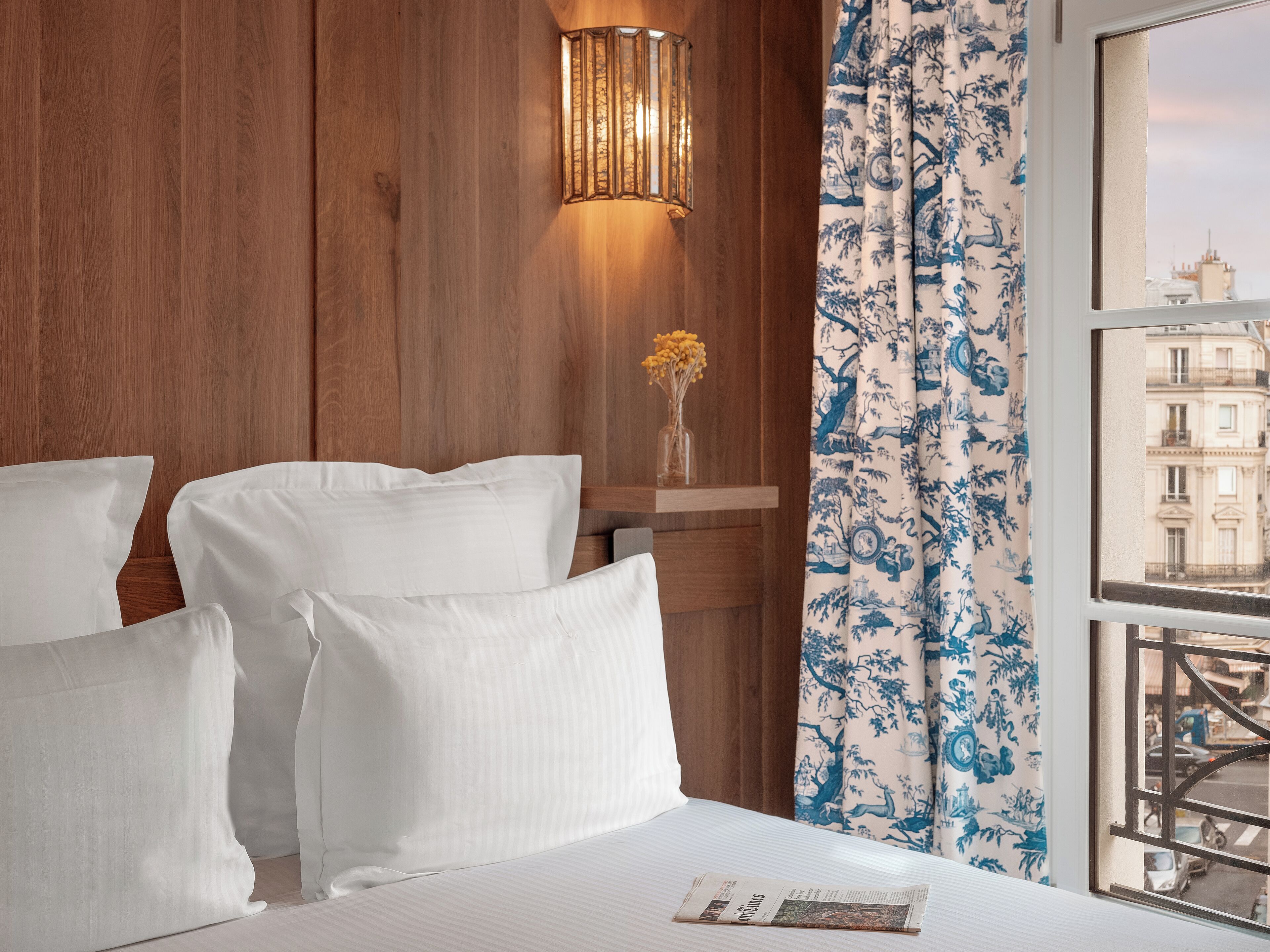 Ein Hotelzimmer mit einer Holzwand, einem Fenster mit blau-blumigen Vorhängen und einem Bett mit weißen Leinen.