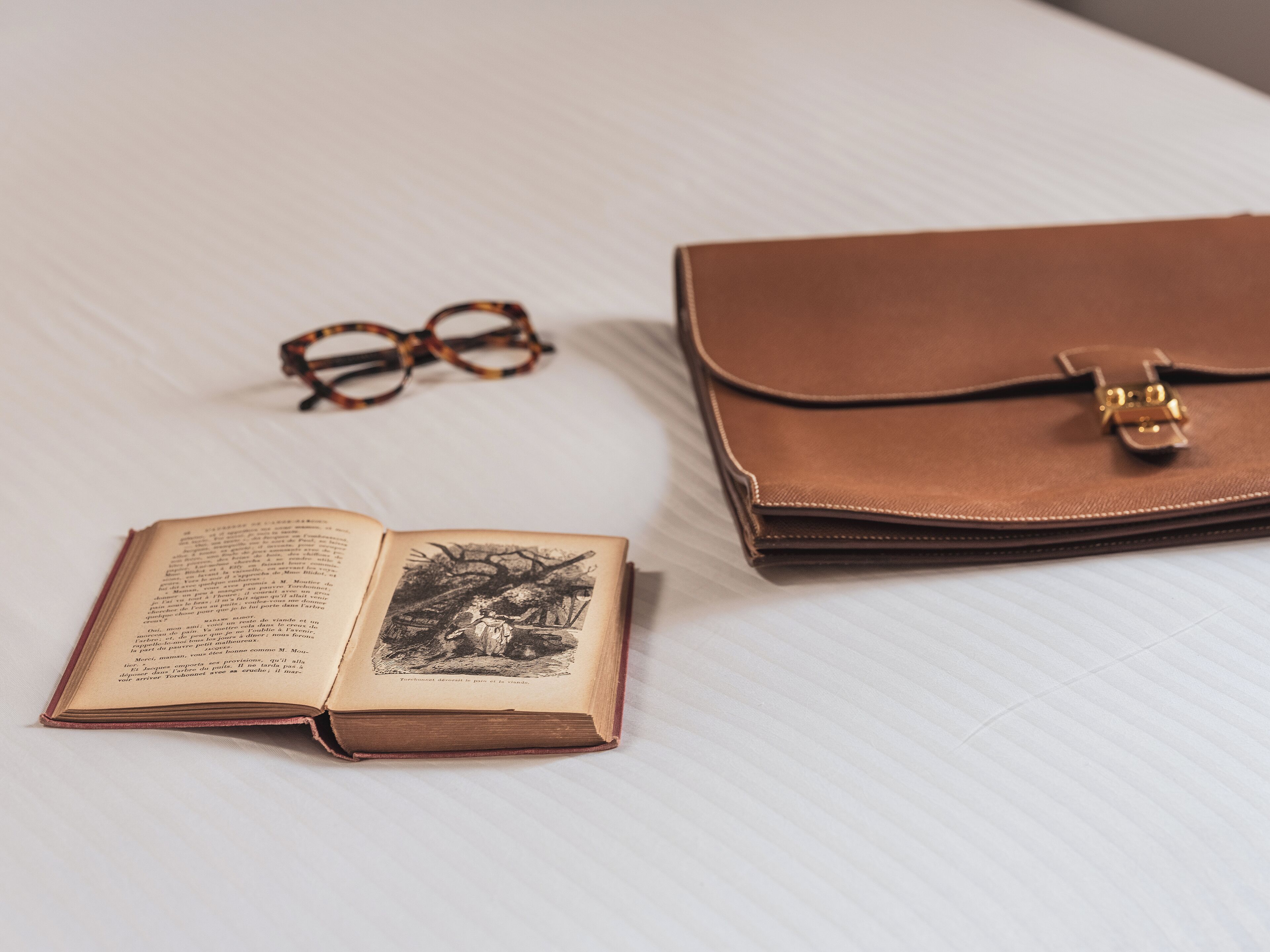 Un libro abierto, gafas y un bolso de cuero marrón cerrado sobre una superficie blanca.