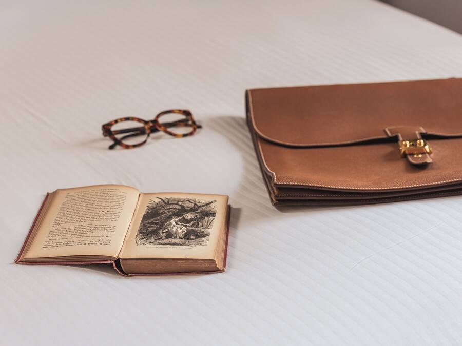 Un libro abierto, gafas y un bolso de cuero marrón cerrado sobre una superficie blanca.