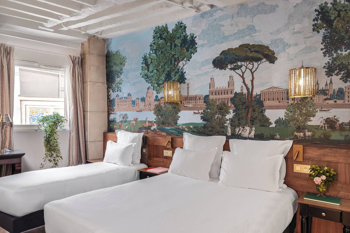 A hotel room with two beds, a mural of a cityscape, a window with curtains, and a nightstand with a lamp and a vase of flowers.
