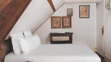 Ein Schlafzimmer mit schrägem Dach, einem Bett mit weißen Bettbezügen, einem Nachttisch mit Buch und Brille, einem kleinen hölzernen Hocker und einem Schreibtisch mit Lampe und gerahmten Bildern an der Wand.