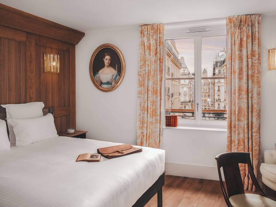 A bedroom with a bed, a nightstand, a chair, a window with curtains, and a framed portrait on the wall.