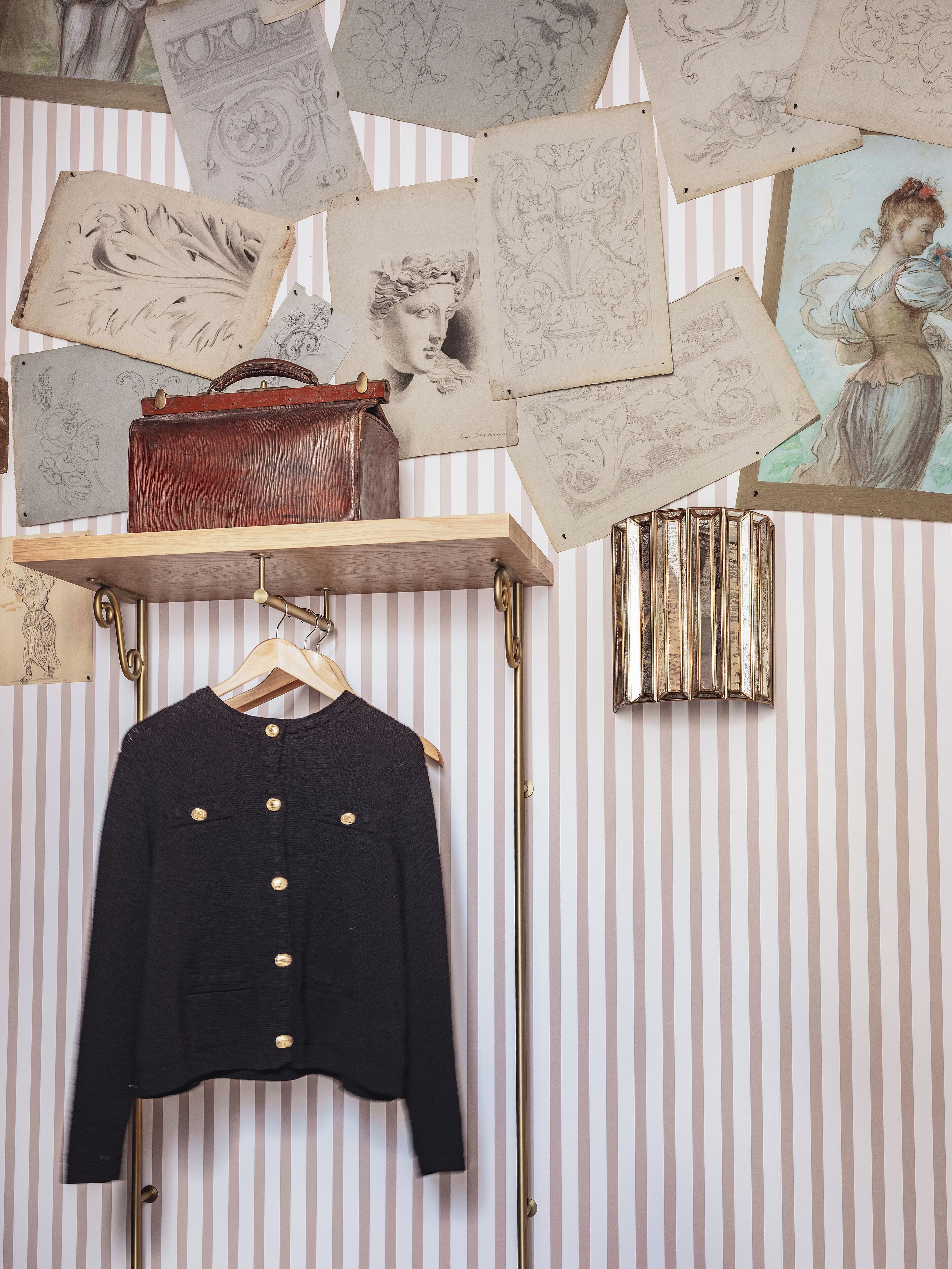 A room with striped wallpaper, a black jacket on a hanger, a red suitcase, and a gold decorative item on the wall.