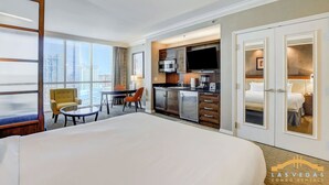 Habitación de hotel con una cama grande, zona de comedor con mesa y sillas, kitchenette y vista a la ciudad.