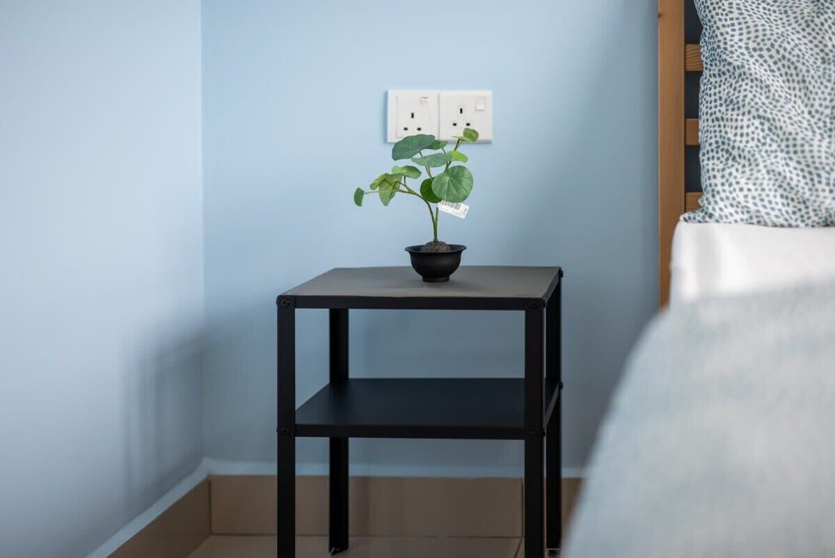 Una mesita metálica negra con una planta en maceta encima, un enchufe blanco en la pared y un cojín estampado en una cama.