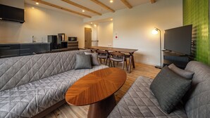 A modern living room with a sofa, a wooden coffee table, a dining table with chairs, a flat-screen TV, and a kitchen area with appliances. - Kanatanosato (Wakayama)