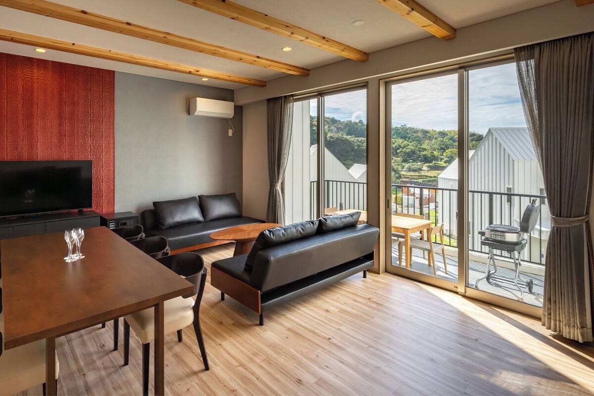 A modern living room with a flat-screen TV, a sofa, a dining table with chairs, and a balcony with a view of the countryside.