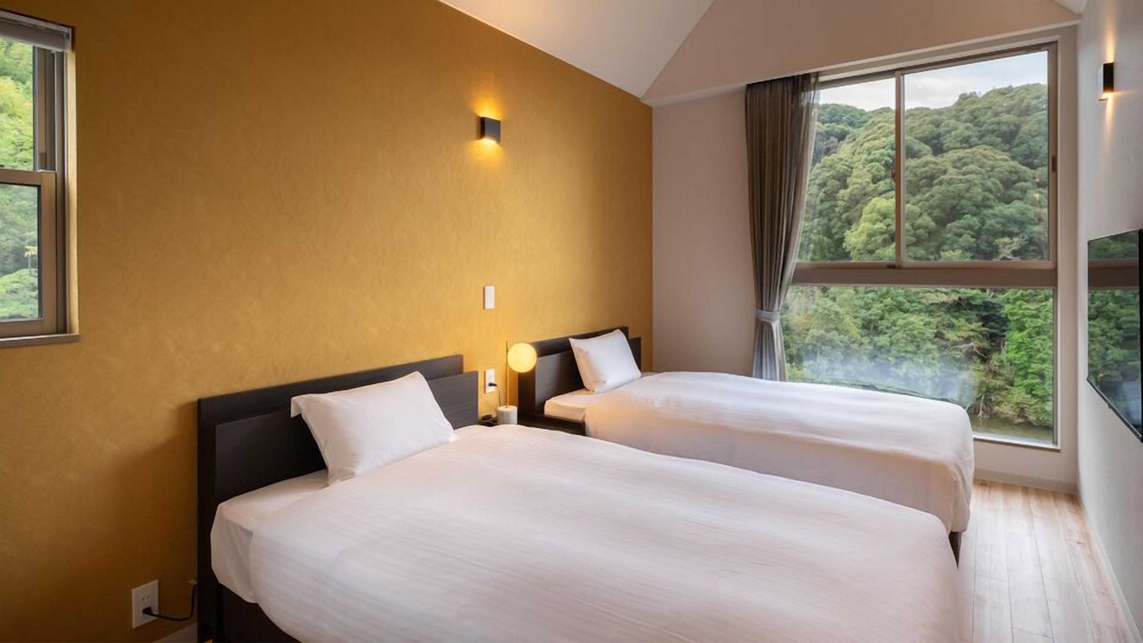 A hotel room with two beds, a large window with a forest view, and a wall-mounted light fixture.