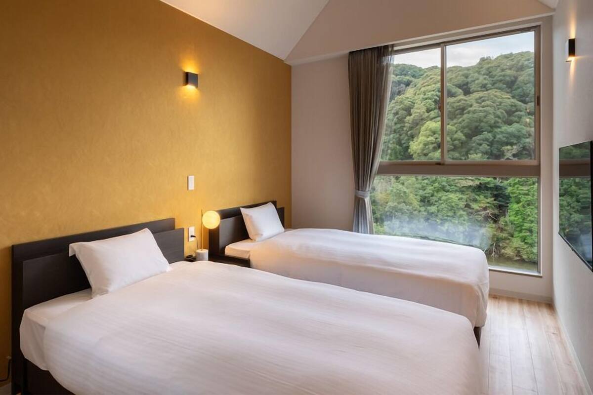 A hotel room with two beds, a large window with a forest view, and a wall-mounted light fixture.