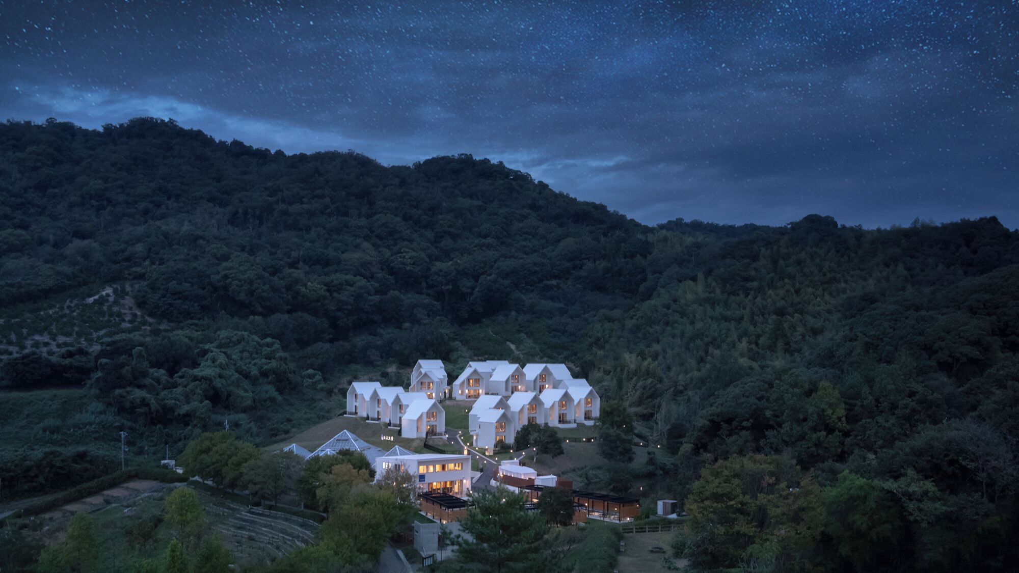 Un conjunto de edificios blancos con numerosas ventanas, rodeado de un bosque denso y colinas, bajo un cielo estrellado.