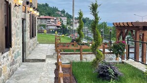 A stone building with a stone pathway leading to a wooden bench area with a view of a town in the background. - Kastel Konak Hotel (Surmene)