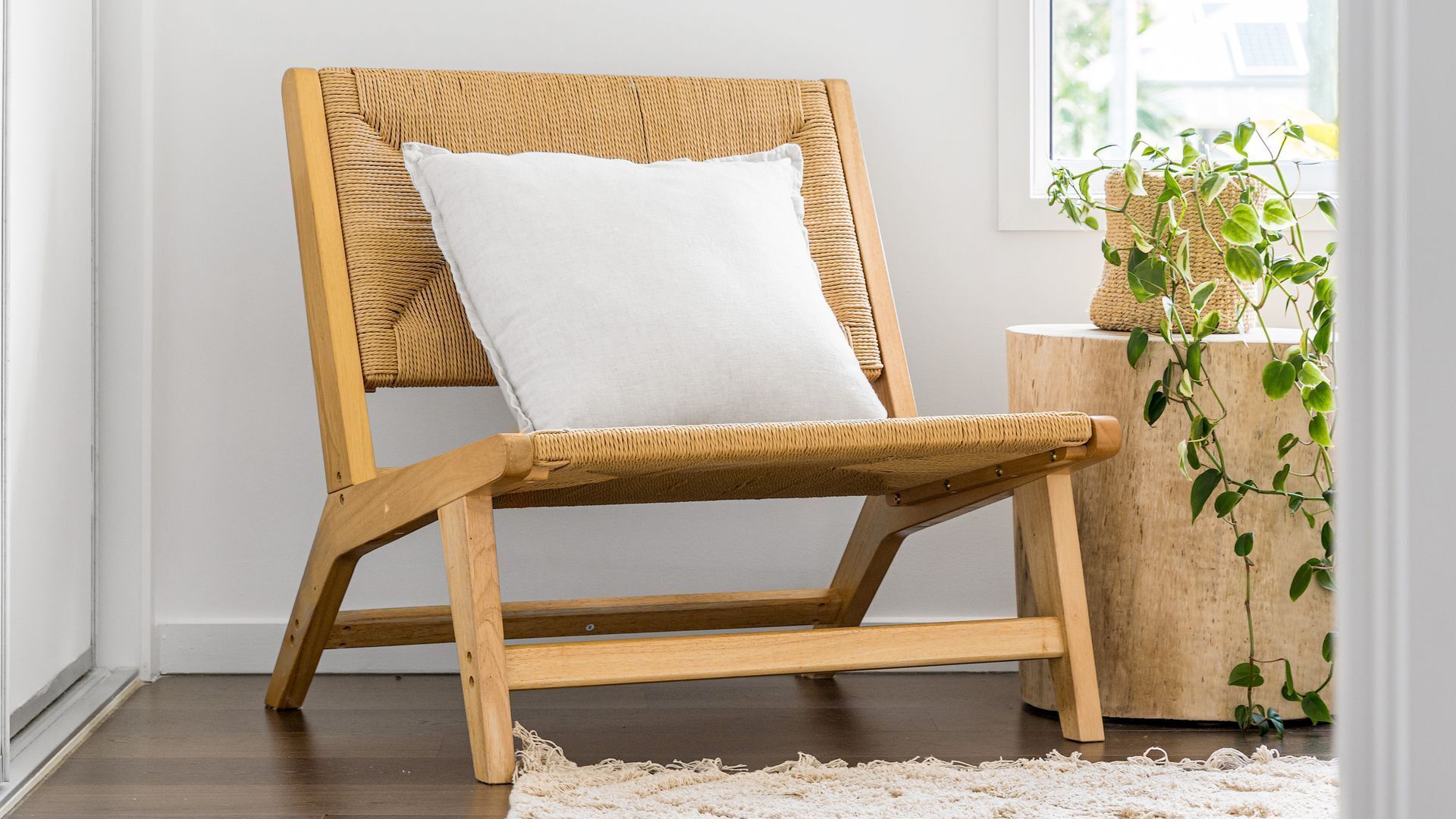 Een rieten stoel met een witte kussens op een kamer met een houten vloer, een klein rond tafeltje en een bloempot.