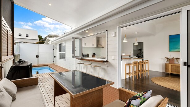 Una cocina moderna con armarios blancos, una zona de bar y un área de estar junto a la piscina con mesa de vidrio y sillas de mimbre.