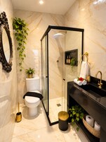 Un baño moderno con ducha envuelta en vidrio, inodoro, lavamanos con detalles dorados y plantas decorativas.