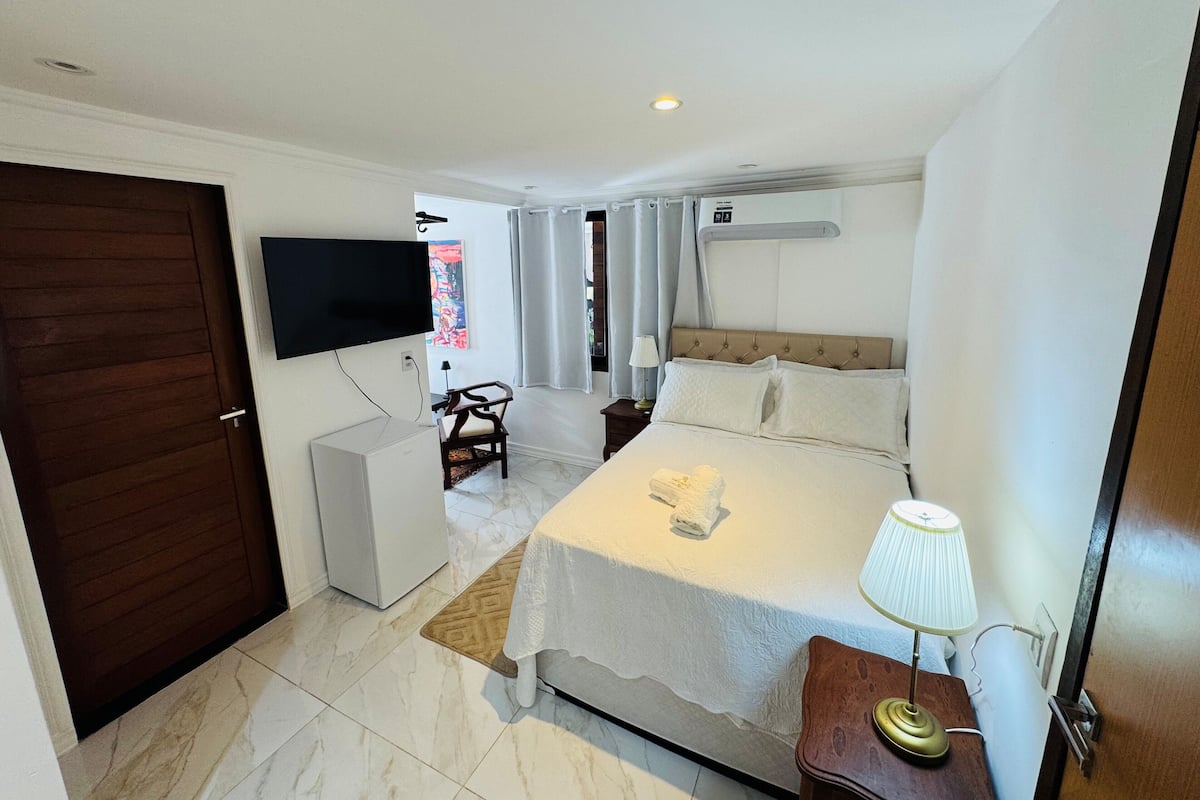 Quarto de hotel com cama, escrivaninha, cadeira, televisão e janela com cortinas.