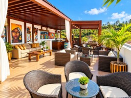Un área de descanso al aire libre con muebles de mimbre, una mesita de centro y una mesa de comedor. Hay plantas en macetas y obras de arte en las paredes.