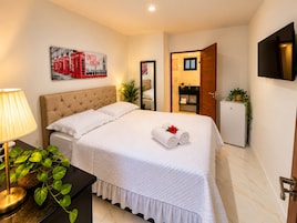 A hotel room with a bed, a bedside table with a lamp, a small refrigerator, a mirror, and a painting of red telephone boxes.