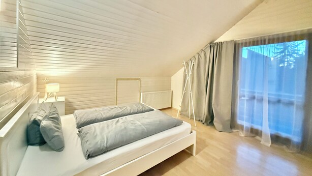 A bedroom with a bed, a nightstand, a lamp, a radiator, and a window with curtains. - H&H HOME - Cosy Rooftop Apartment with Loggia (Sindelfingen)