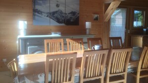 A wooden cabin interior with a dining table, chairs, a kitchen area, and a mountain landscape painting on the wall.