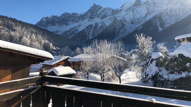 A wooden chalet with snow-covered roofs, set against a backdrop of snow-capped mountains and clear blue sky.