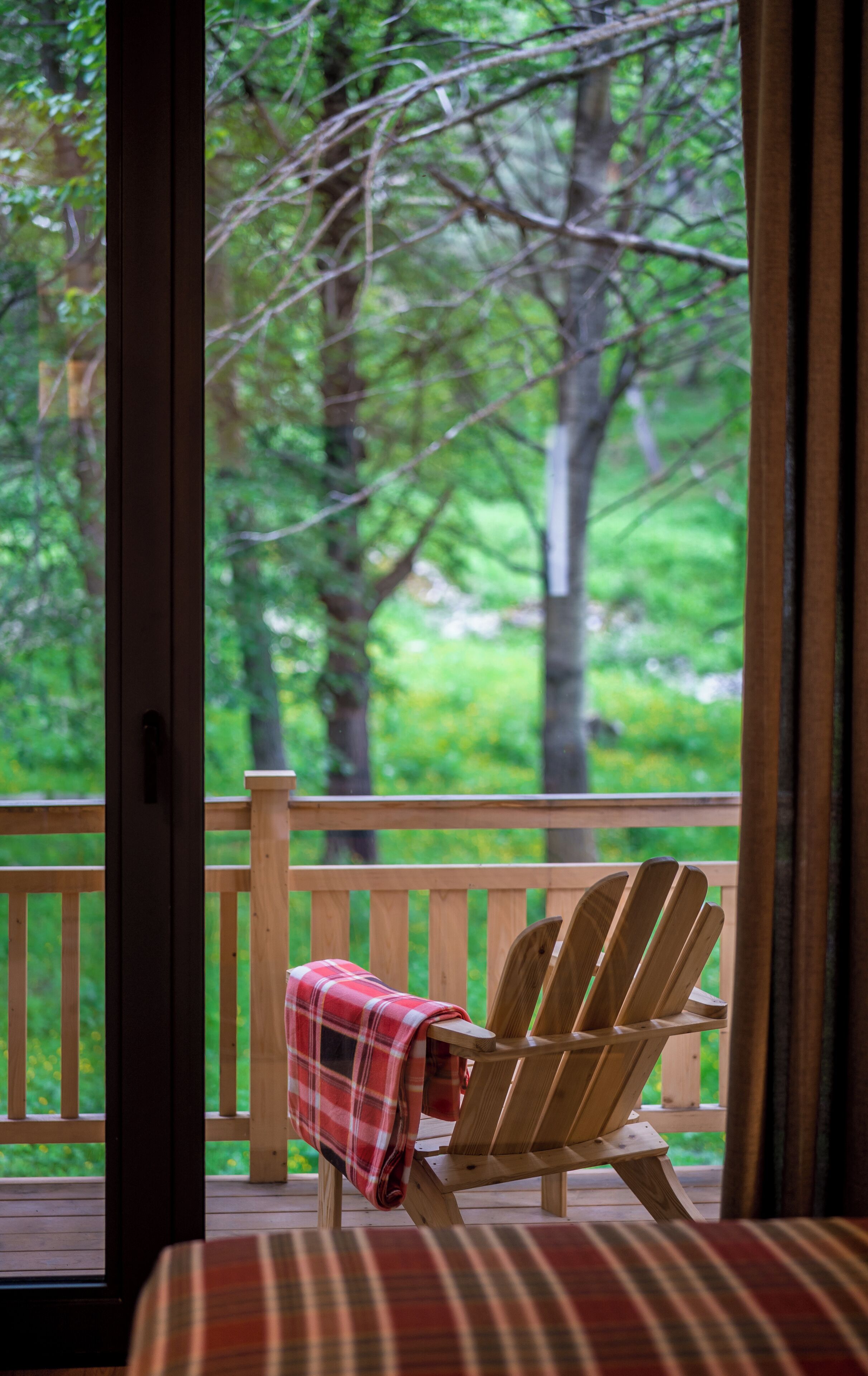 A wooden deck with a plaid blanket on a wooden Adirondack chair, overlooking a forested area.