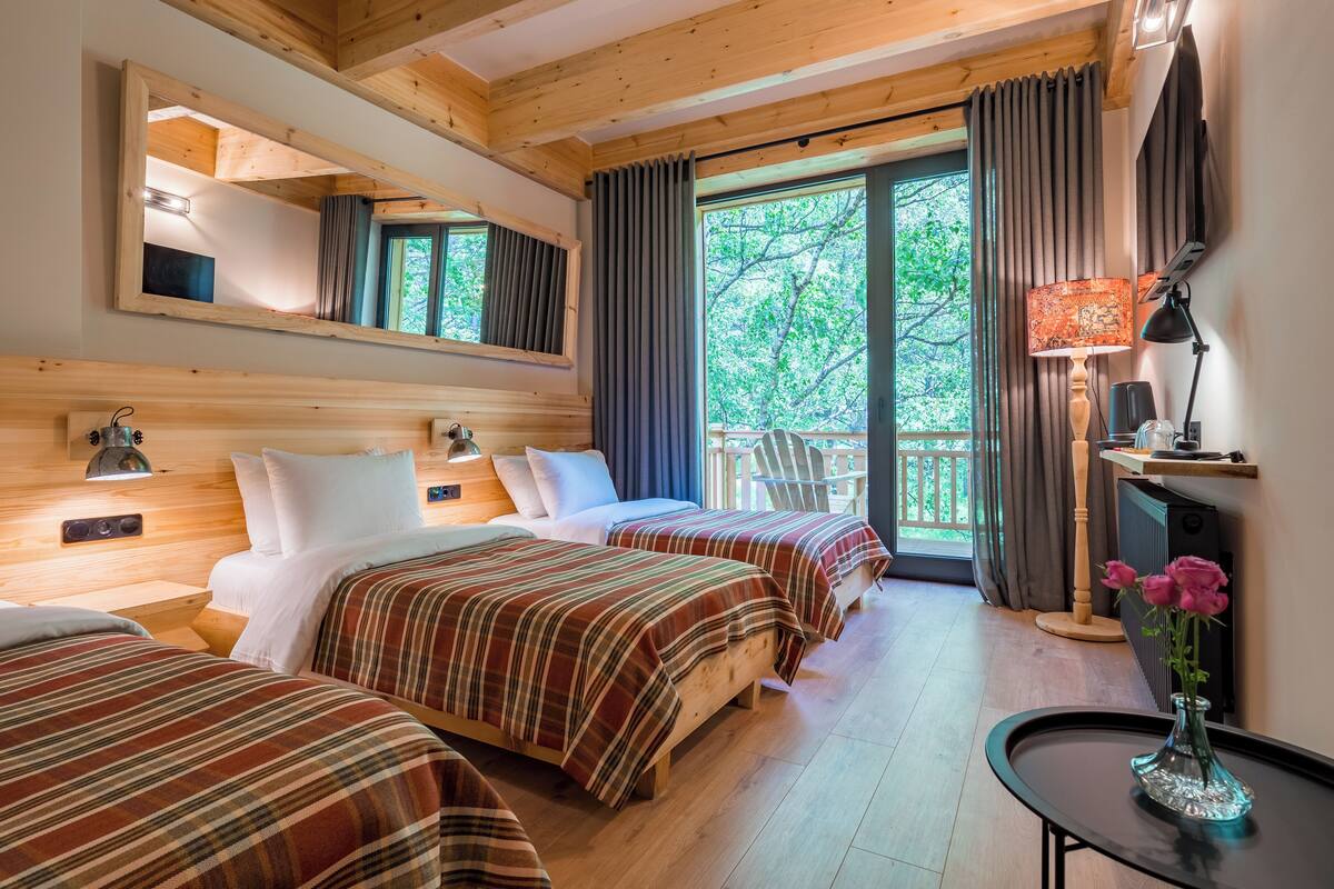 A hotel room with two beds, a wooden ceiling, a large window with curtains, a small table with a vase, and a chair by the window.