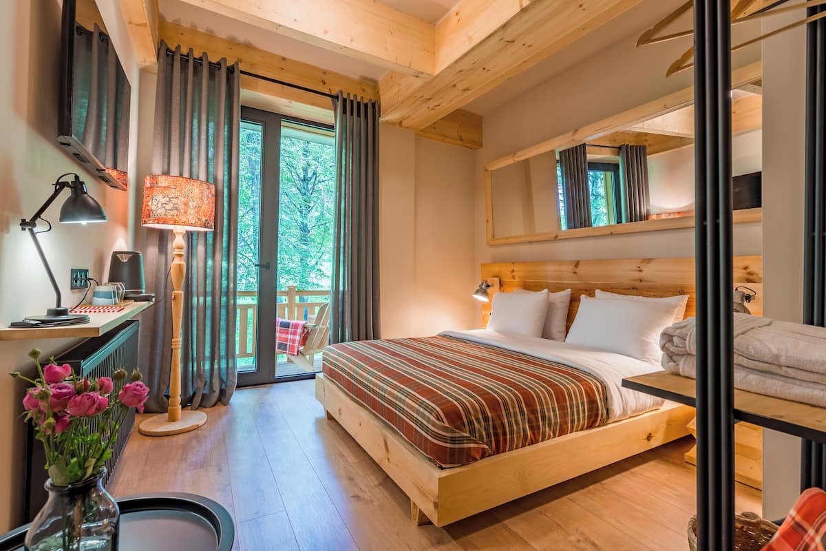 A bedroom with a wooden bed, a desk with a lamp, a chair, and a view of greenery outside.