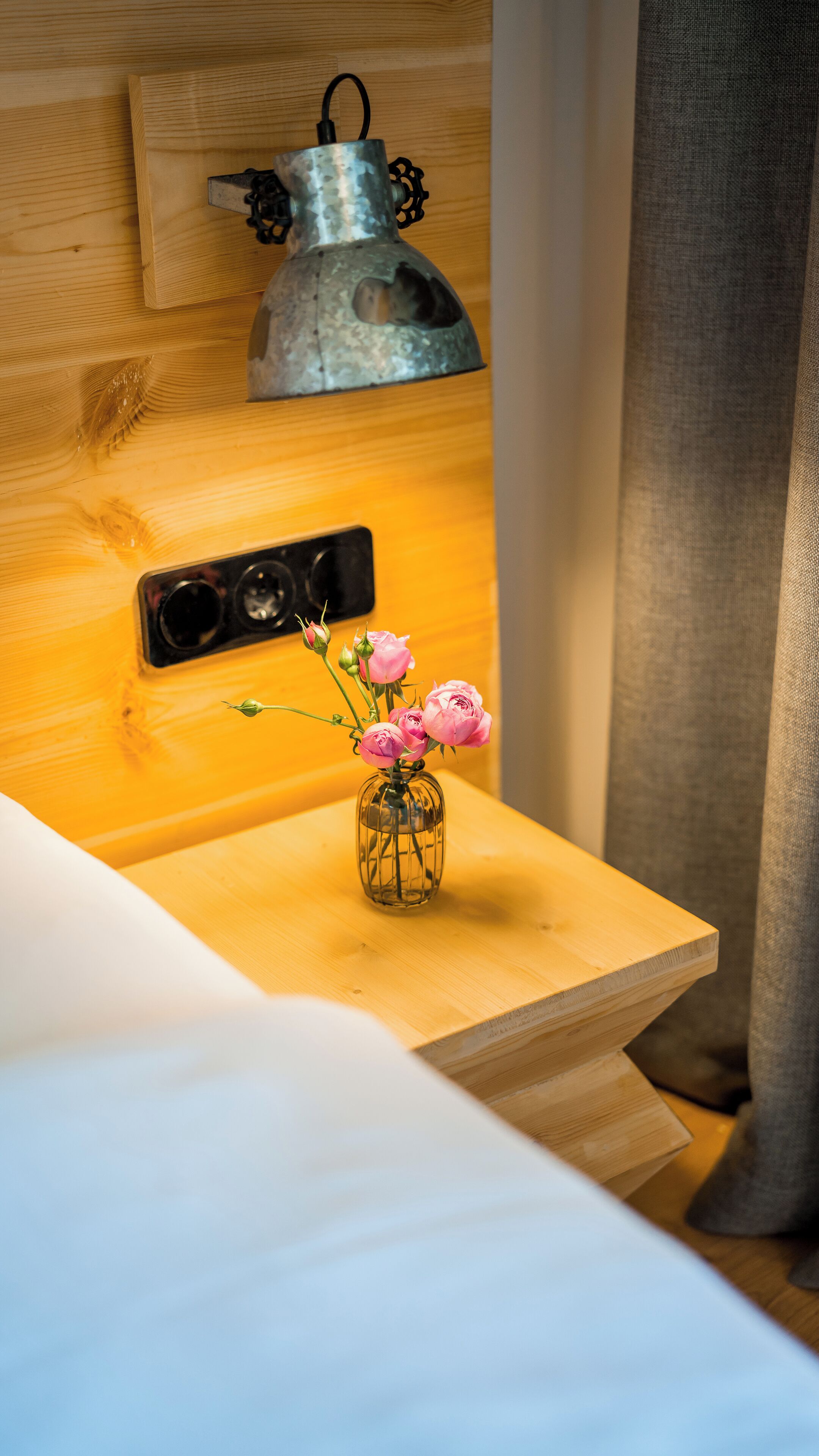 A wooden bedside table with a clear glass vase containing pink flowers, a metal lamp, and a power outlet.