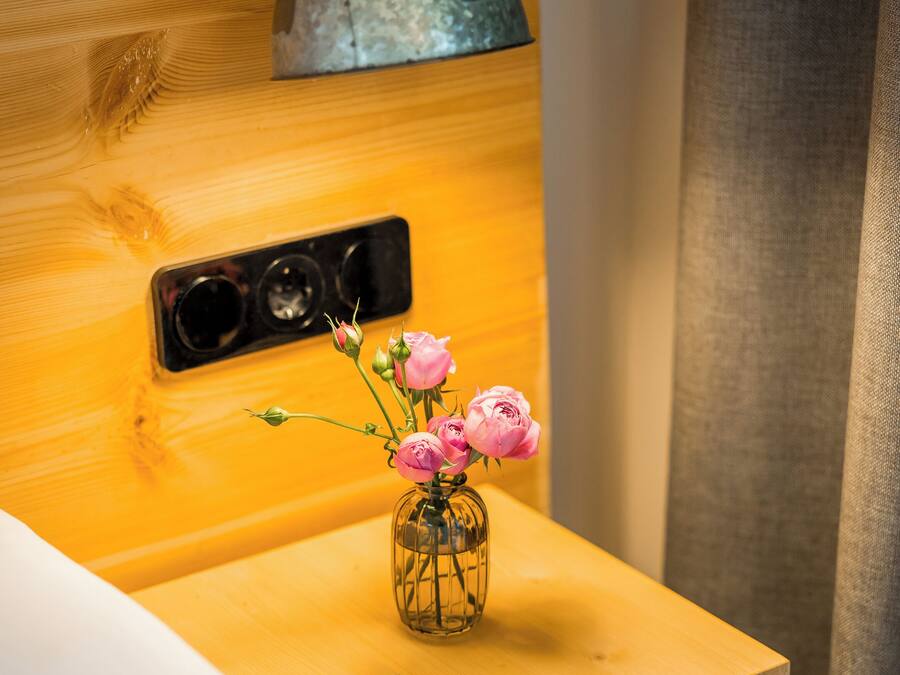 A wooden bedside table with a clear glass vase containing pink flowers, a metal lamp, and a power outlet.