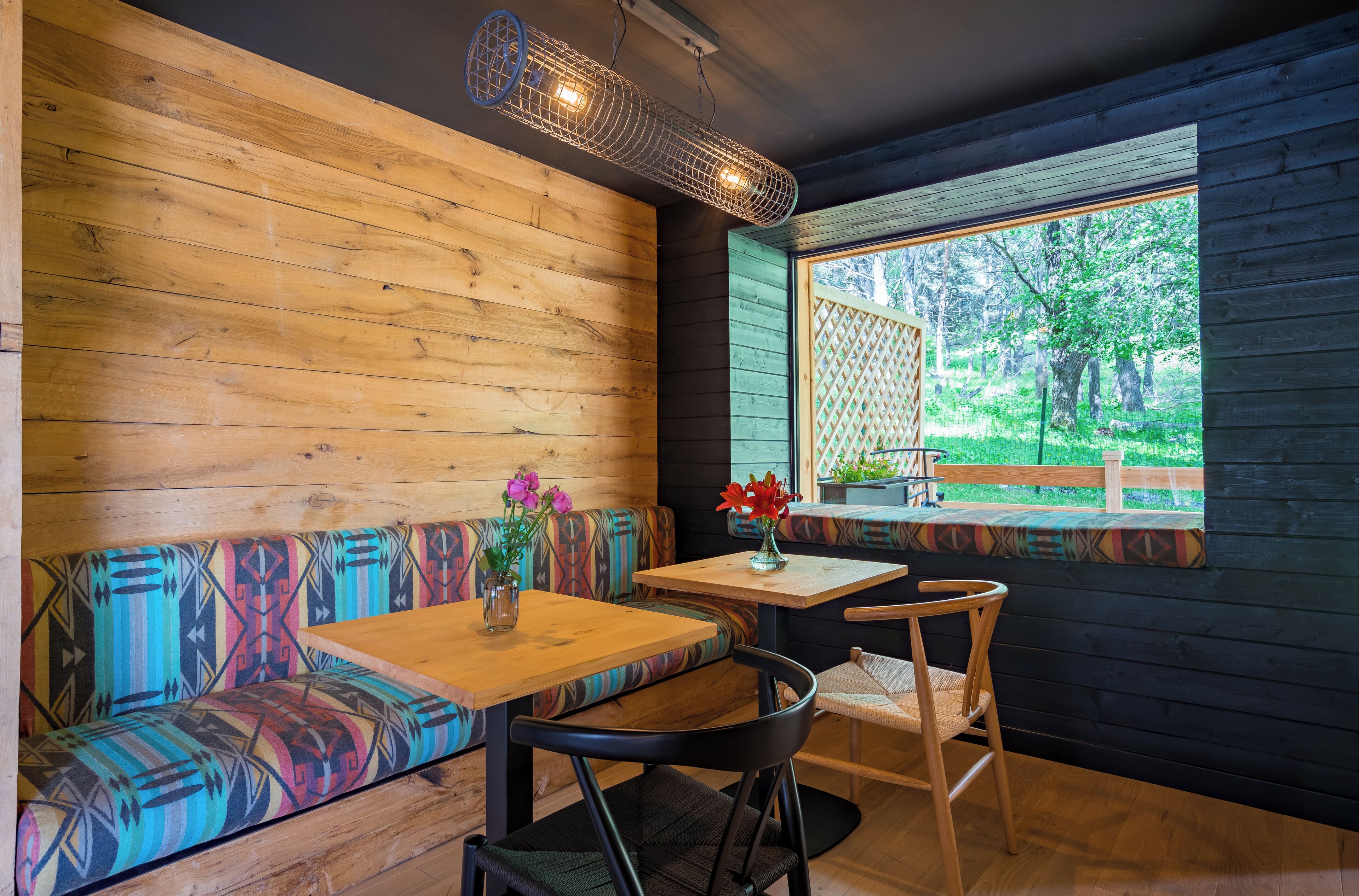 A wooden interior with a bench, table, and chairs, a window with a view of greenery, and a hanging light fixture.