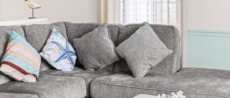 A living room with a grey sectional sofa, a white coffee table, and a window with sheer curtains.