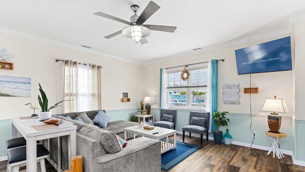 A living room with a grey sofa, a white coffee table, a ceiling fan, and a TV mounted on the wall. - Downtown OC Townhome! - (B) (Ocean City)