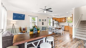 A spacious living area with a wooden dining table, white bar stools, a ceiling fan, and a kitchen in the background. - Downtown OC Townhome! (A) (Ocean City)
