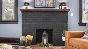 A modern living room with a dark brick fireplace, a framed picture above it, and a potted plant on the right. - Walk to State Street - Remodeled Home (Bristol)
