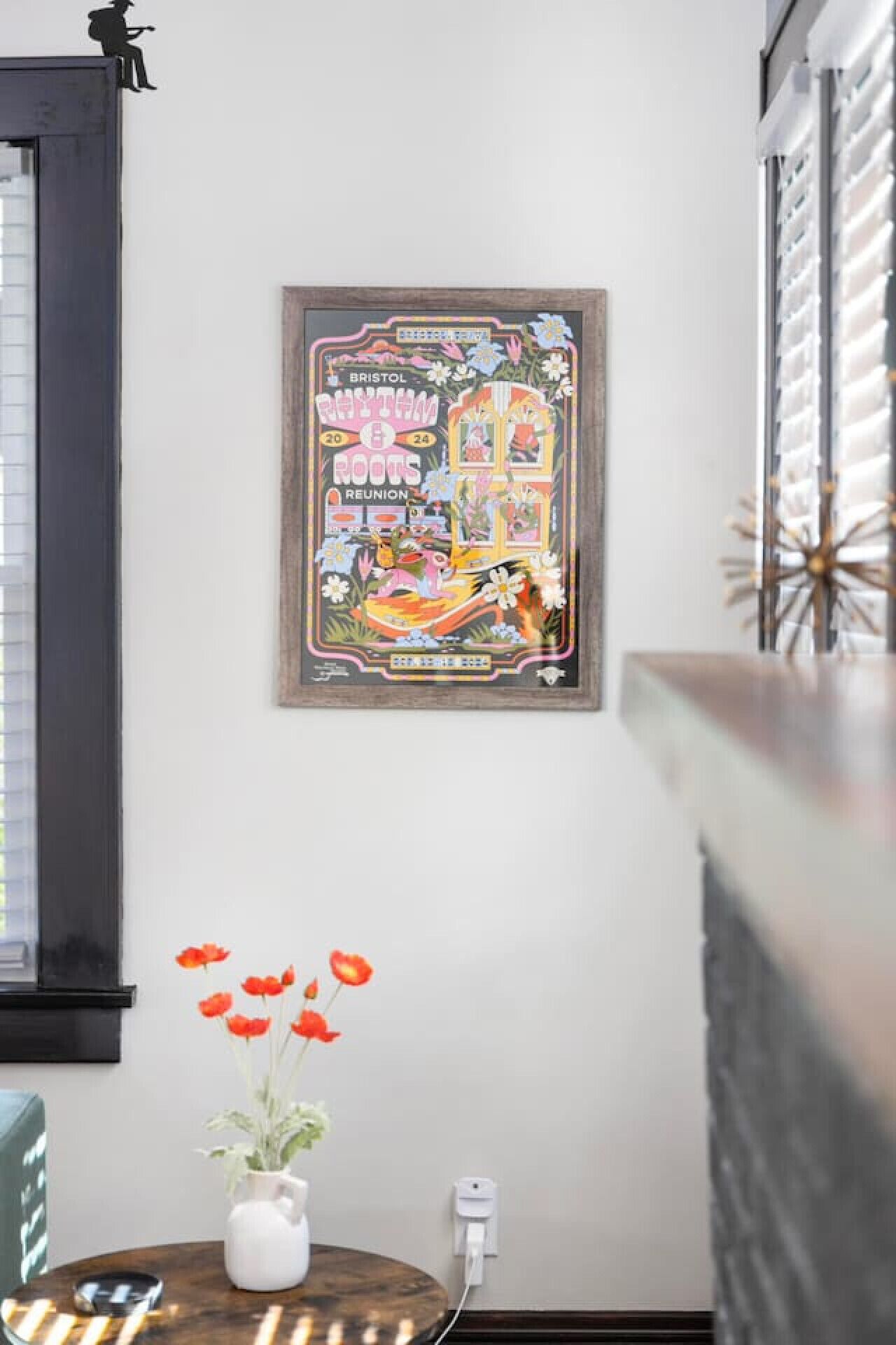 Et indrammet plakat på væggen, et lille bord med en vase blomster, og en stikkontakt.