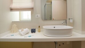 Ein Badezimmer mit weißem Waschbecken, einem Spiegel, einem Fenster mit Jalousie und einer Duschkabine.