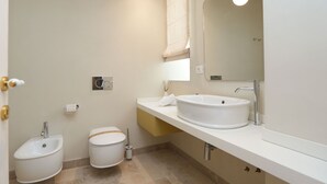 Ein modernes Badezimmer mit einem weißen Waschbecken, einem Bidet und einer Toilette. Über dem Waschbecken hängt ein Spiegel, darüber eine Leuchte.