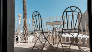 Ein Balkon mit zwei Metallstühlen und einem kleinen Tisch, mit Blick auf einen Strand mit Sonnenschirmen und Palmen.